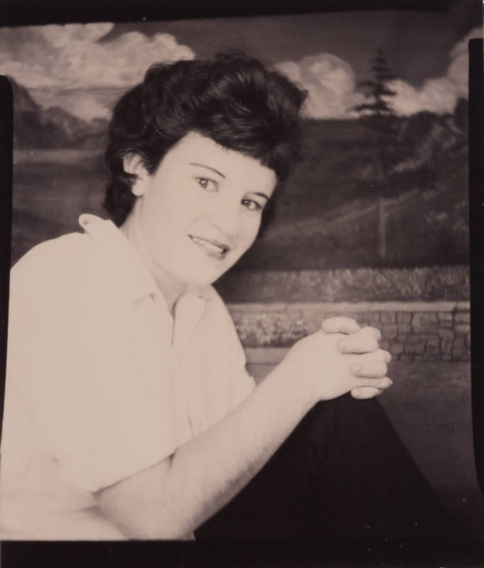 Photo booth portrait of a woman with dark hair and clasped hands resting on her knees