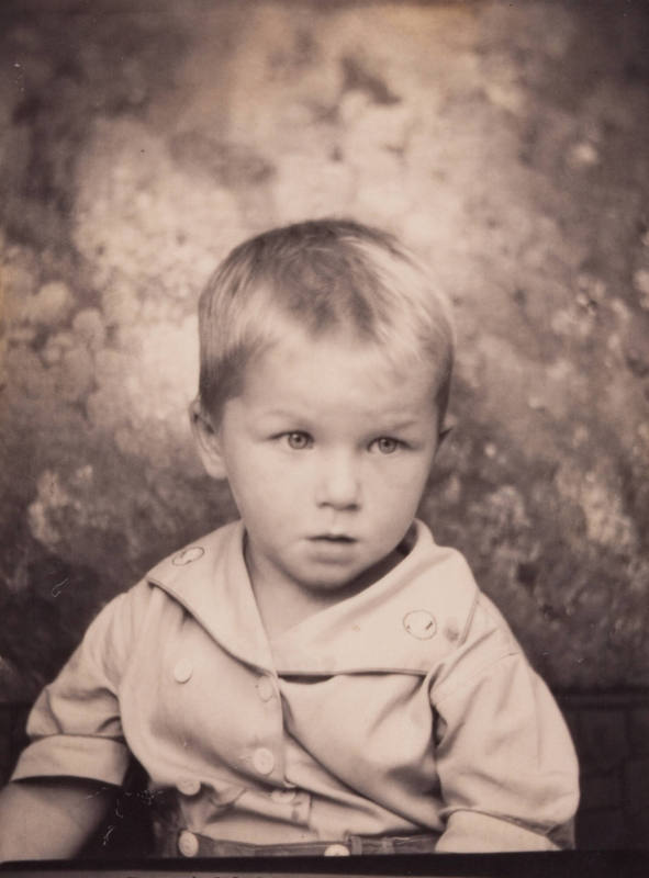 Photo booth portrait of a little boy with light-colored hair combed forward, wearing a shirt with a large collar and buttons