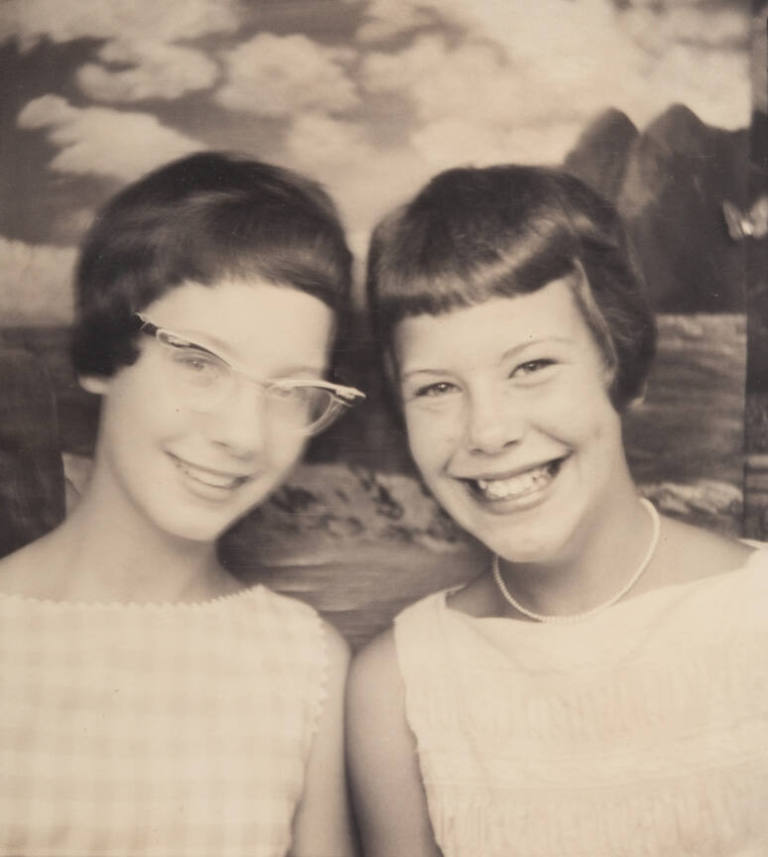 Photo booth portrait of two young girls, possibly sisters or twins, one wearing cat eye glasses and the other wearing a choker-style necklace