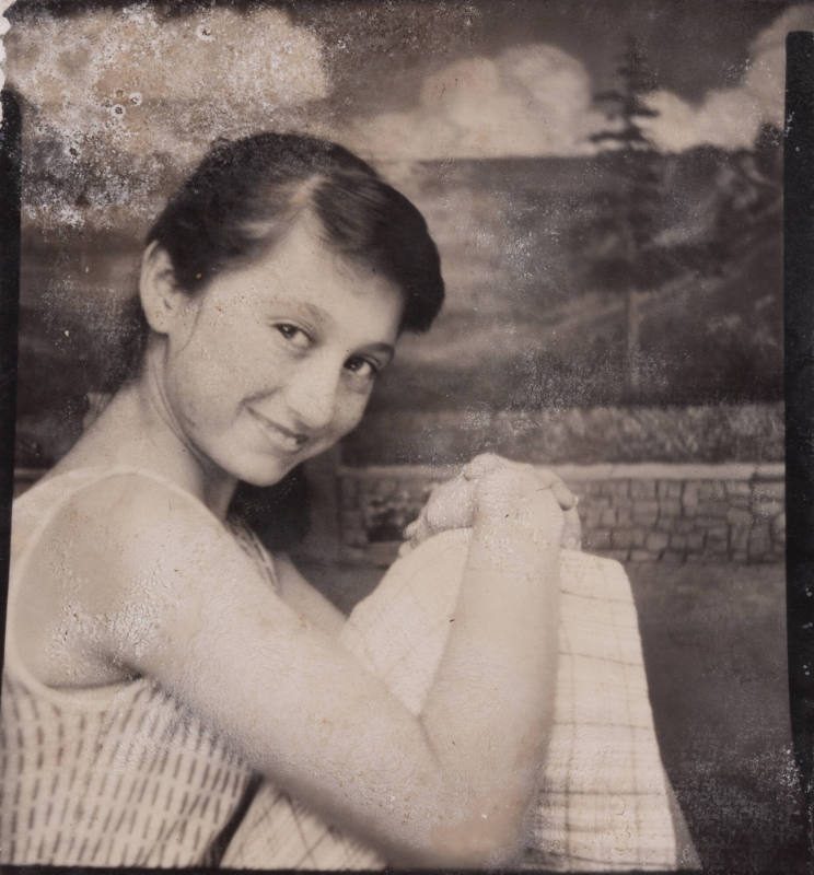 Photo booth portrait of a girl or teen with her knees drawn up and hands clasped, wearing a sleeveless top and grid patterned skirt