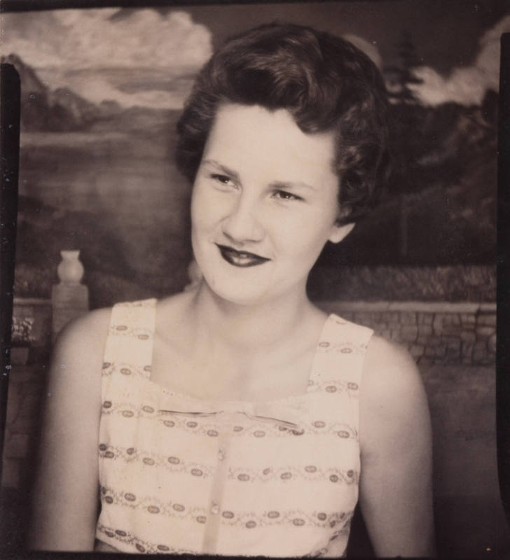 Photo booth portrait of a young woman with dark hair, wearing lipstick, a sleeveless dress, and smiling with mouth closed