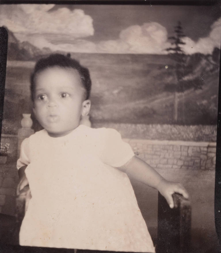Photo booth portrrait of a baby or toddler, partly seated, gripping the armrest of his chair or stool