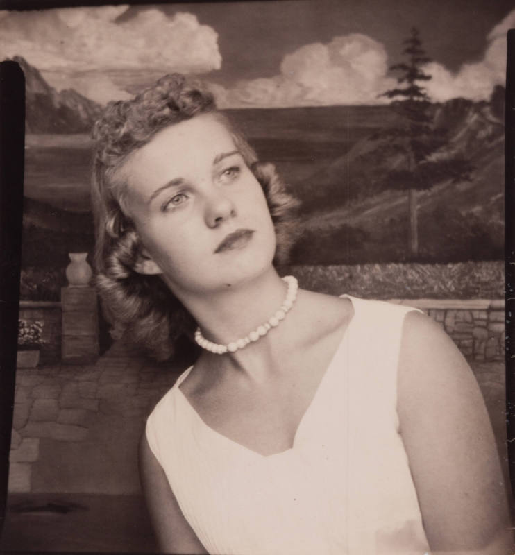 Photo booth portrait of a young woman with curly, light-colored hair, wearing a choker necklace high on her neck, and posed in a leaning, tilted position