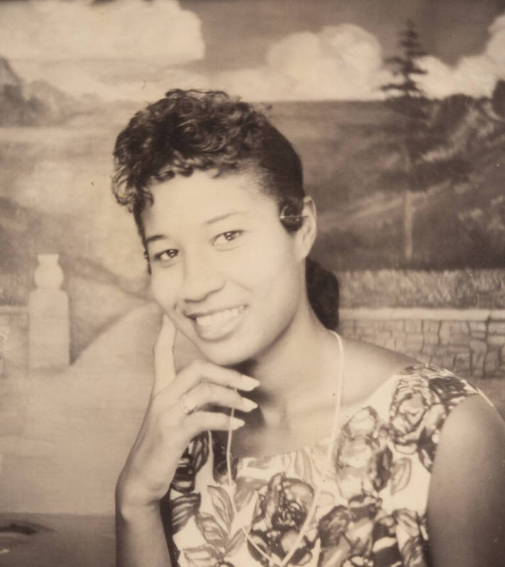 Photo booth portrait of a young woman wearing a floral dress or top, posed with her hand to her chin and one finger on her cheek