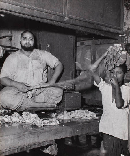 [Butcher brandishing a switch over cuts of meat to keep the flies away in his stall at the market, Lahore, India]