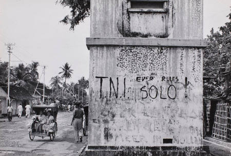 Two women in a rickshaw pedaling past graffitied cement block, Java