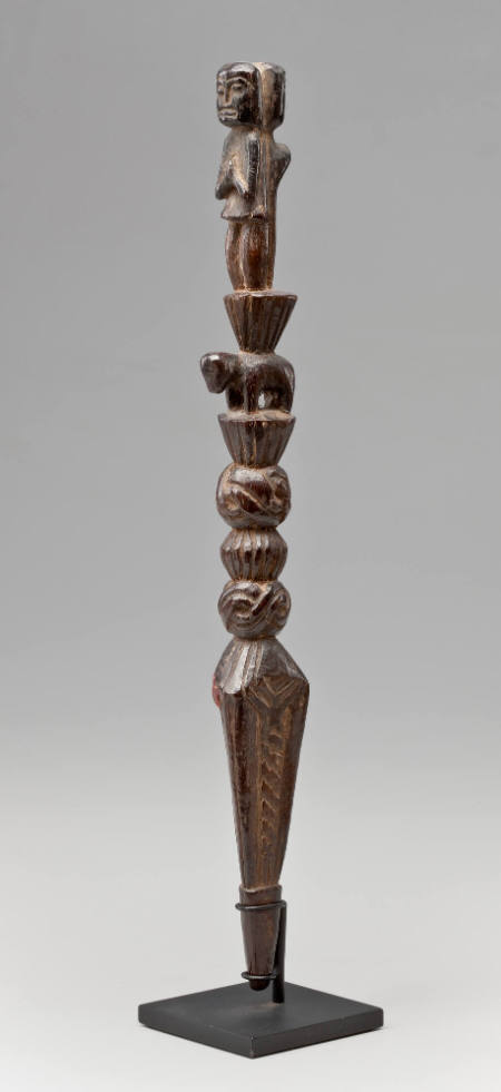 Wooden staff with two figures at top, one with "praying" hands, the other behind a snake, standing on animal's back